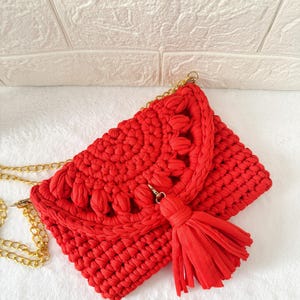 May include: A vibrant red crochet clutch with a gold chain strap and a matching tassel. The bag features a textured design with decorative elements along the flap. The clutch is set against a white background.