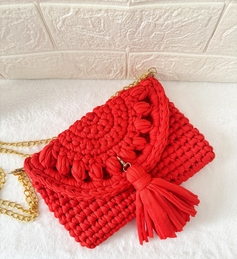 May include: A vibrant red crochet clutch with a gold chain strap and a matching tassel. The bag features a textured design with decorative elements along the flap. The clutch is set against a white background.