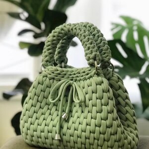 May include: A sage green handbag with a woven texture. The bag features two rounded handles and a drawstring closure with silver-toned metal accents. The bag is made of a soft, thick material.