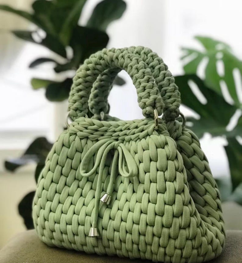 May include: A sage green handbag with a woven texture. The bag features two rounded handles and a drawstring closure with silver-toned metal accents. The bag is made of a soft, thick material.