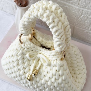 May include: Cream-colored, hand-knitted handbag with a rounded shape and a thick, braided handle. The bag features gold-colored metal rings and drawstring closure with gold-colored accents. The bag is made of a soft, chunky knit material.