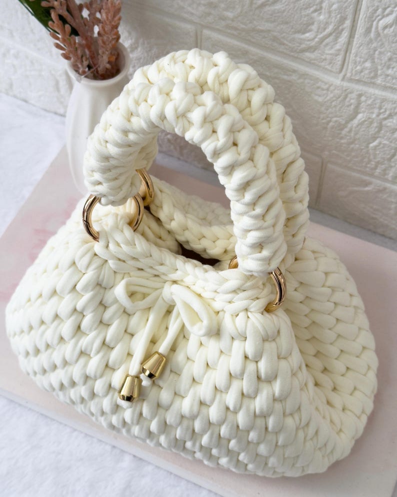 May include: Cream-colored, hand-knitted handbag with a rounded shape and a thick, braided handle. The bag features gold-colored metal rings and drawstring closure with gold-colored accents. The bag is made of a soft, chunky knit material.