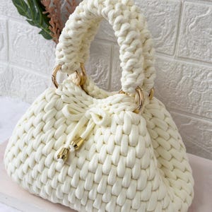 May include: Cream-colored, hand-knitted handbag with a woven texture. The bag features a rounded shape, a drawstring closure, and gold-colored metal rings connecting the handles. The handles are thick and looped.