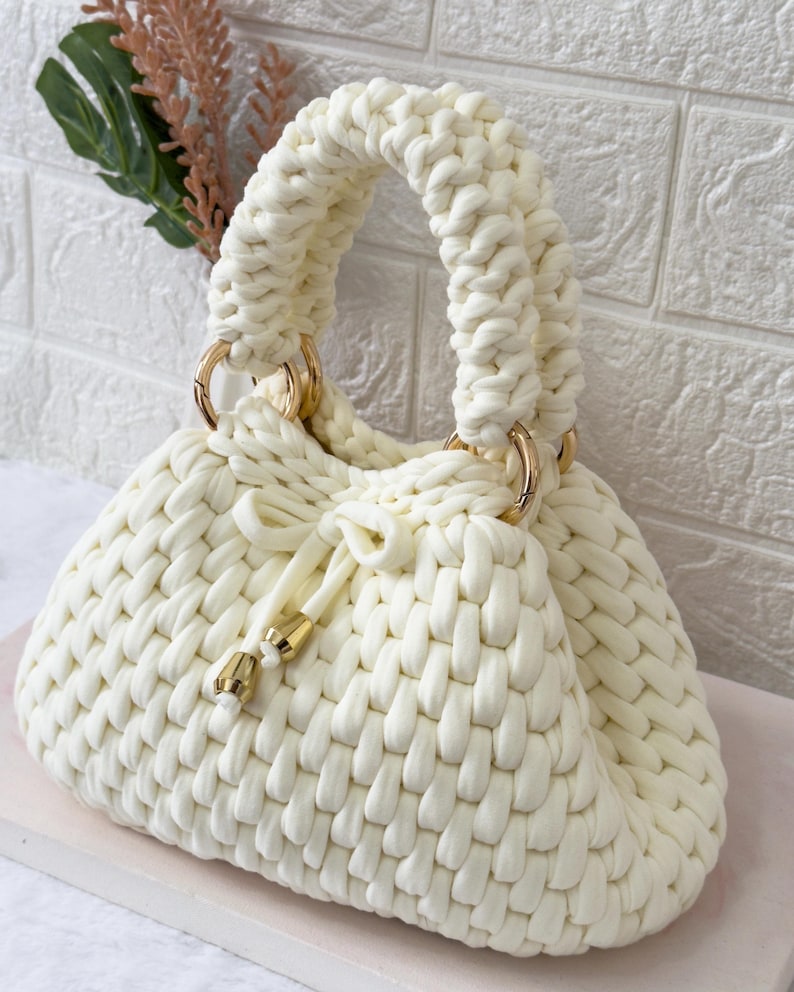May include: Cream-colored, hand-knitted handbag with a woven texture. The bag features a rounded shape, a drawstring closure, and gold-colored metal rings connecting the handles. The handles are thick and looped.