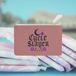 May include: A dusty rose-colored zippered pouch with the words "Cycle Slayer" in black gothic font, a crescent moon, and a purple teardrop. Two purple roses are at the bottom. The pouch is on top of a stack of feminine hygiene products.