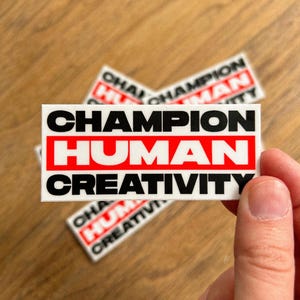 May include: Rectangular stickers with the words "CHAMPION HUMAN CREATIVITY" in bold black font, with "HUMAN" in a red rectangle. Several stickers are visible, with one held by a hand. The stickers are on a wooden surface.