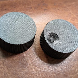 May include: Two dark gray, cylindrical knobs with ridged sides and flat tops. One knob has a circular indentation on top. The knobs are made of a textured material and sit on a brown surface.