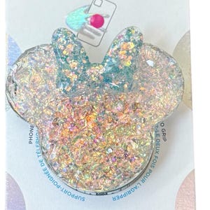 Op de afbeelding: Een witte PopGrip telefoonhouder met een glitterontwerp in de vorm van Mickey Mouse's hoofd. De houder is verpakt op een witte kaart met het PopSockets-logo en de tekst "PopGrip".