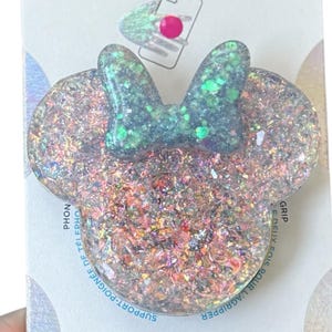 Op de afbeelding: Een witte en groene glitter telefoonhouder met een roze strik en de tekst "POPGRIP" op de verpakking.