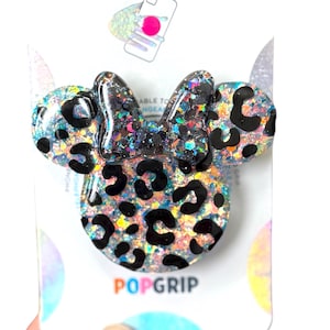 Op de afbeelding: Een Minnie Mouse-vormig PopGrip-telefoonaccessoire. Het accessoire heeft een glinsterende, iriserende basis met zwarte luipaardvlekken. De strik en oren zijn ook glinsterend. Het woord "PopGrip" staat in oranje onderaan.