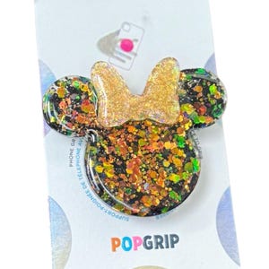 Op de afbeelding: Een Minnie Mouse-vormige telefoonhouder met een gouden glitterstrik en zwarte basis. De basis is gevuld met kleurrijke glitter. De telefoonhouder is bevestigd aan een witte achterkant met de tekst "POPGRIP".