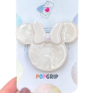 Puede incluir: Un PopSockets PopGrip blanco con forma de cabeza de ratón con un lazo. El PopGrip está en una tarjeta blanca e iridiscente con las palabras "POPSOCKETS" y "POPGrip" impresas.