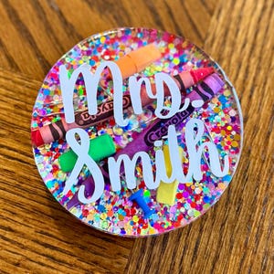 May include: A round, glitter-filled resin coaster with the words "Mrs. Smith" in white script. The coaster features colorful confetti and miniature crayons, a push pin, and a yellow eraser. The design is a vibrant mix of colors, perfect for a teacher.