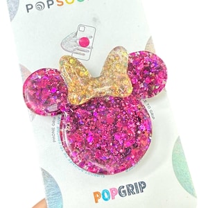 May include: A pink glitter PopSocket shaped like a cartoon character's head with a gold glitter bow. The PopSocket is on a white card with the words "POPSOCKET" and "POPGRI" printed on it.