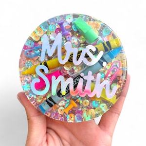 May include: A round, clear resin coaster with colorful sequins and school supplies. The coaster features a yellow crayon, blue crayon, pink paperclip, and the text "Mrs. Smith" in a holographic font. A push pin and a green highlighter are also visible.