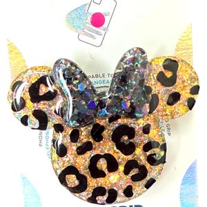 Op de afbeelding: Een PopGrip telefoonaccessoire in de vorm van een muizenkop met een strik. Het accessoire heeft een luipaardprint ontwerp met zwarte vlekken en gouden glitter. De strik is zwart met iriserende glitter. Het woord "POPGRIP" is in oranje gedrukt.