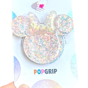 Op de afbeelding: Een doorzichtige, met glitter gevulde PopSockets PopGrip in de vorm van Minnie Mouses hoofd en strik. De PopGrip is bevestigd aan een witte en lichtblauwe PopSockets-merkverpakking. Het woord "POPSOCKETS" staat bovenaan de kaart.
