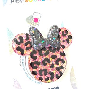 Op de afbeelding: Een PopSockets PopGrip met een Minnie Mouse-ontwerp. De telefoonhouder heeft een roze en zwarte luipaardprint met een zwarte glitterstrik. De verpakking leest "PopSockets" en "PopGrip".