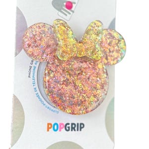 Op de afbeelding: Een glitterende telefoonhouder in de vorm van een muizenkop met een strik. De houder is voornamelijk roze met gouden glitteraccenten. De verpakking leest "POPGRIP" en "PHONE GRIP".