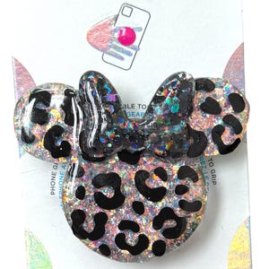 Op de afbeelding: Een PopGrip-telefoonaccessoire met een Minnie Mouse-ontwerp. De grip heeft een luipaardprintpatroon met zwarte vlekken en glitteraccenten. De strik is zwart met glitter. Het is bevestigd aan een witte kaart.