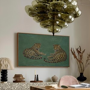 May include: An interior design scene featuring a wooden desk, a pink office chair, and two leopard-print ottomans. A painting of two leopards hangs above a cabinet with a black and white spotted design. A large, green glass chandelier is suspended from the ceiling.