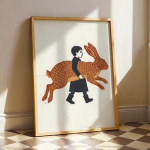 May include: A framed art print featuring a stylized illustration of a person with dark hair wearing a black coat and boots, carrying a large, textured orange-brown rabbit. The artwork is set against a light, off-white background with subtle flecks.