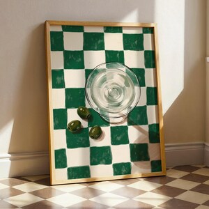 May include: A framed art print featuring a green and white checkered pattern. A clear glass martini glass and three green olives are on the print. The frame is a light wood color.