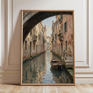 May include: A framed painting depicting a narrow canal in Venice, Italy. The scene features weathered multi-story buildings in shades of beige and pink, a small wooden boat floating on the water, and a stone archway framing the top of the view.