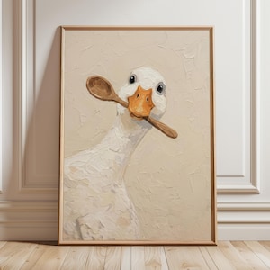 Op de afbeelding: Een ingelijste kunstdruk met een schilderij van een witte gans die een houten lepel in zijn bek houdt. De gans heeft een feloranje snavel en staat tegen een getextureerde, gebroken witte achtergrond. Het kunstwerk is ingelijst in een lichtbruine houten lijst.
