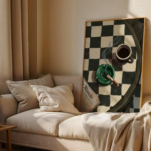 May include: A framed artwork depicts a still life with a cup of coffee, a lit cigar in a green ashtray, and a newspaper. The background features a black and white checkered pattern. The artwork is displayed above a beige sofa.