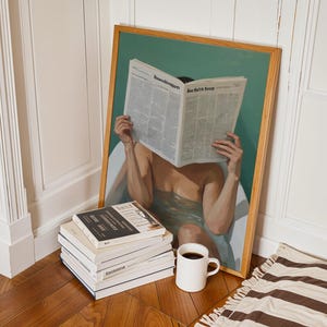 Könnte beinhalten: Ein gerahmtes Kunstwerk zeigt eine Person in einer Badewanne, die eine Zeitung liest. Ein Stapel Bücher, eine weiße Tasse mit Kaffee und ein gestreifter Teppich liegen auf dem Holzboden. Das Kunstwerk hat einen Holzrahmen und einen grünen Hintergrund.