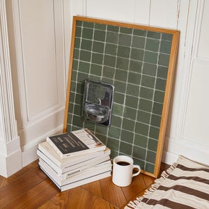 以下が含まれることがあります： 緑色のタイル張りの壁のアートワークが額装され、金属製の石鹸ディスペンサーが壁に立てかけられています。 本の山、白いコーヒーマグ、ストライプのラグが床にあります。 フレームは木製です。