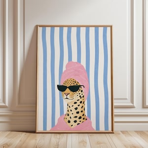 May include: A framed art print featuring a leopard wearing sunglasses, a pink towel on its head, and a pink robe. The background has blue and white vertical stripes. The artwork is displayed on a wooden floor.