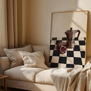 May include: A framed still life painting featuring a dark brown stovetop espresso maker, figs, and a black and white checkered pattern. The painting is displayed on a beige wall above a beige sofa.