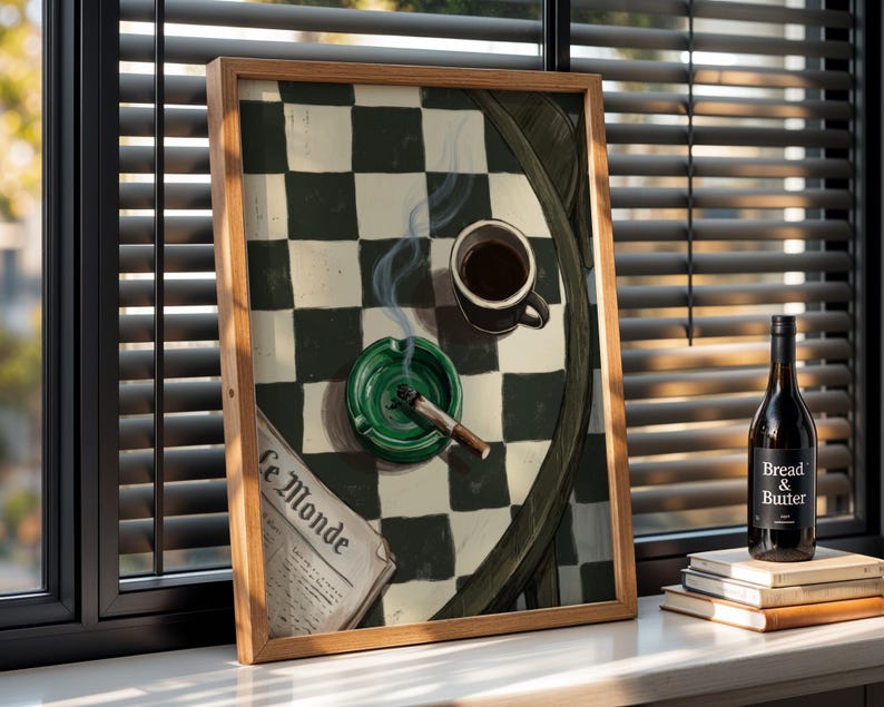 May include: A framed artwork depicts a still life with a cup of coffee, a lit cigarette in a green ashtray, and a newspaper. A bottle of wine and a stack of books are in the background. The artwork is set against a window with blinds.
