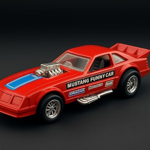 May include: A red toy Mustang Funny Car with a blue stripe on the hood. The car has a large engine, a rear spoiler, and the words "MUSTANG FUNNY CAR" on the side. The wheels are silver.