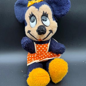 May include: A vintage plush toy of a cartoon mouse wearing a blue dress with orange and white polka dots and a yellow bow. The mouse has large, black eyes and a wide smile.