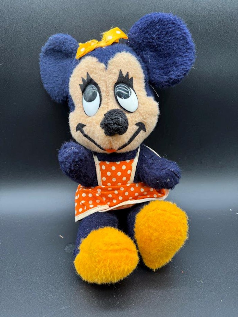 May include: A vintage plush toy of a cartoon mouse wearing a blue dress with orange and white polka dots and a yellow bow. The mouse has large, black eyes and a wide smile.