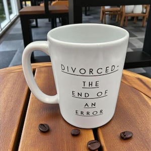 May include: White ceramic mug with the text "DIVORCED - THE END OF AN ERROR" in black. The mug sits on a wooden surface with scattered coffee beans. The mug has a curved handle.