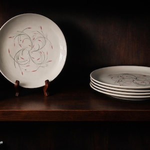 Peut inclure: Assortiment d'assiettes en céramique blanche avec un motif floral gris et rose. Une assiette est présentée sur un présentoir en bois. D'autres assiettes sont empilées sur une étagère en bois foncé, à côté d'un vase rose et d'une tasse grise foncée.