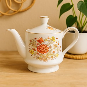 May include: A white ceramic teapot with a lid, featuring a floral design in shades of orange, yellow, and gold. The teapot has a gold trim and a curved handle and spout. A decorative piece for serving tea.