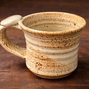 May include: Handmade ceramic mug with a unique handle design. The mug features a speckled, earthy color palette with shades of beige and brown. The cylindrical body has a textured surface, perfect for enjoying coffee or tea.