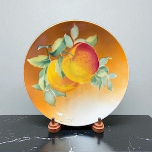 May include: A decorative plate with a hand-painted design of two apples on a branch with green leaves. The plate has an orange and white gradient background and is displayed on a wooden stand.