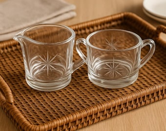 Op de afbeelding: Twee heldere glazen room- en suikerset met sterrenpatronen, geplaatst op een bruin geweven dienblad. De creamer heeft een tuit en een handvat, de suikerpot heeft twee handvatten. Ideaal voor het serveren van koffie of thee.