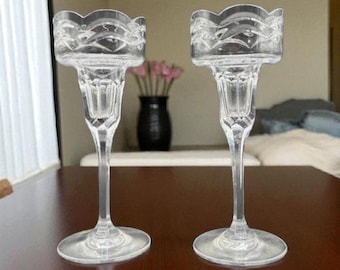 Könnte beinhalten: Zwei klare Kerzenhalter aus Glas mit dekorativem, gewelltem Rand. Die Kerzenhalter haben einen schlanken Stiel und einen runden Fuß. Ein dunkelbrauner Tisch befindet sich im Vordergrund, und eine Vase mit rosa Blumen im Hintergrund.