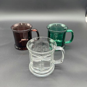Puede incluir: Tres tazas de vidrio con asas. Una es transparente, otra es marrón oscuro y otra es verde. La taza transparente está en primer plano, con las tazas marrón y verde detrás. Todas las tazas tienen un diseño similar.