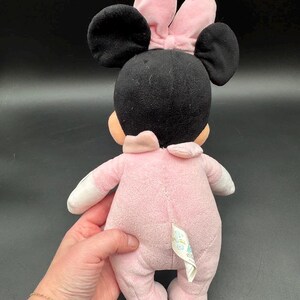May include: A plush Minnie Mouse doll, viewed from the back, in a pink onesie with white accents. The doll has black ears and a pink bow. The doll is being held by a person.