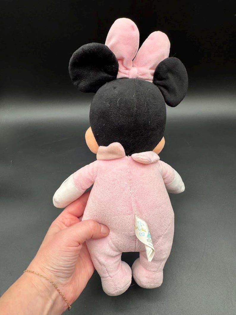 May include: A plush Minnie Mouse doll, viewed from the back, in a pink onesie with white accents. The doll has black ears and a pink bow. The doll is being held by a person.