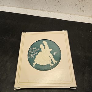 Peut inclure: Une boule de Noël verte et blanche avec une silhouette d'un couple patineur sur glace. La boule porte l'inscription "First Christmas Together" et "1982".