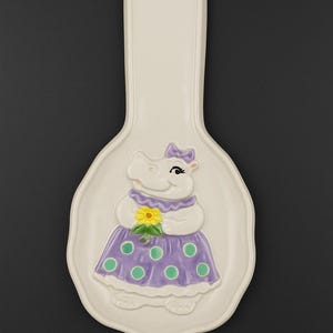 Op de afbeelding: Crèmekleurige keramische lepelsteun met een nijlpaardontwerp. Het nijlpaard draagt een paarse jurk met groene stippen en houdt een gele bloem vast. De lepelsteun heeft een handvat met een gat om op te hangen.